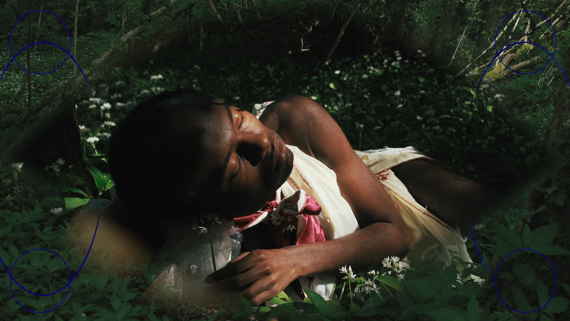  A close up of a person lying on the ground with their eyes closed. They're in a dark woodland under a small patch of sunlight. They're wearing white and surrounded by wild garlic flowers.