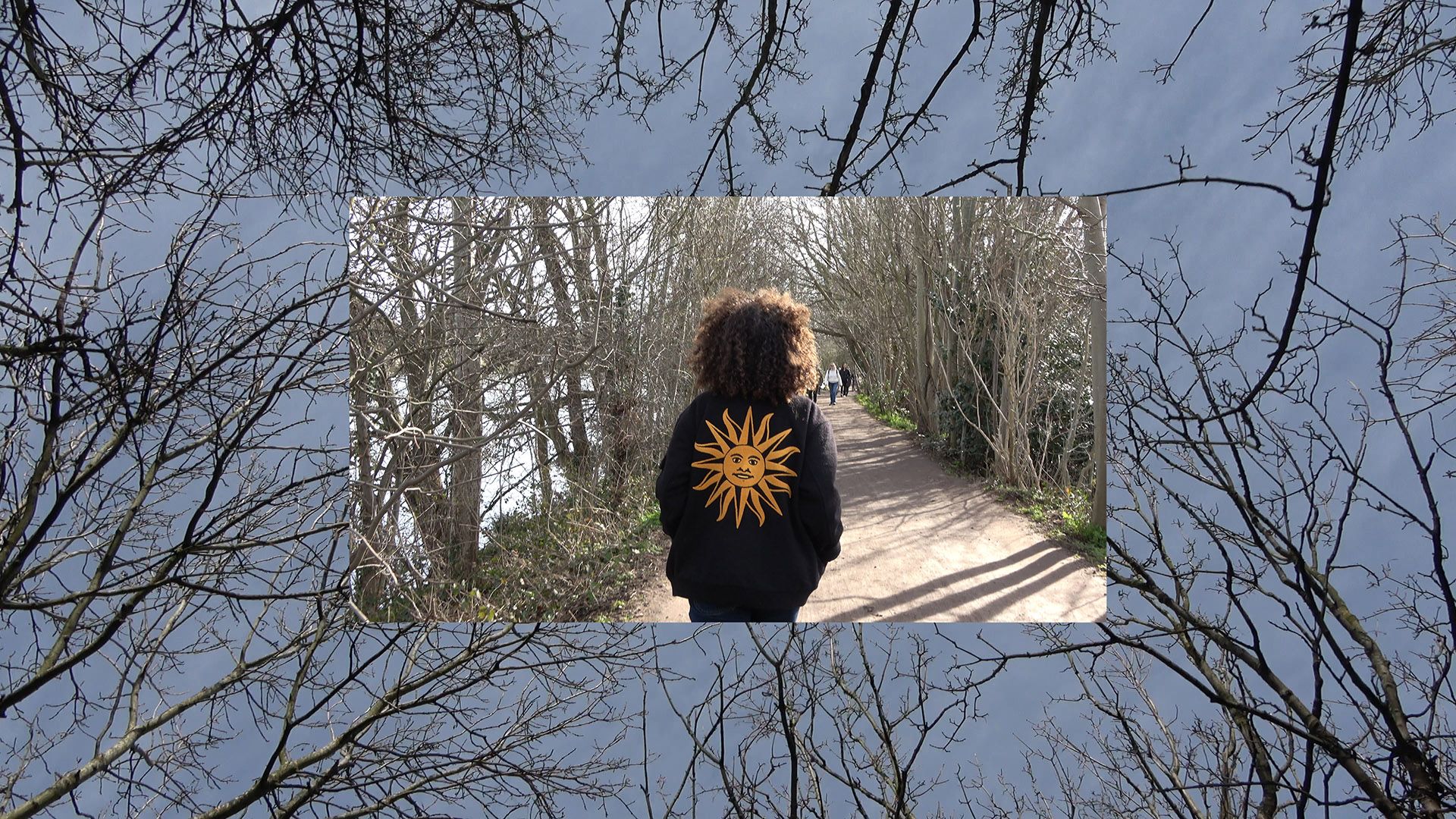 An image of Jamila from the back. She's walking down a pedestrian path lined by bare trees and a river on the left. Jamila is wearing a black jacket which has an image of a yellow sun with a face on the back. The image is overlaid on a larger image of blue sky with bare tree branches coming into the frame from all sides.